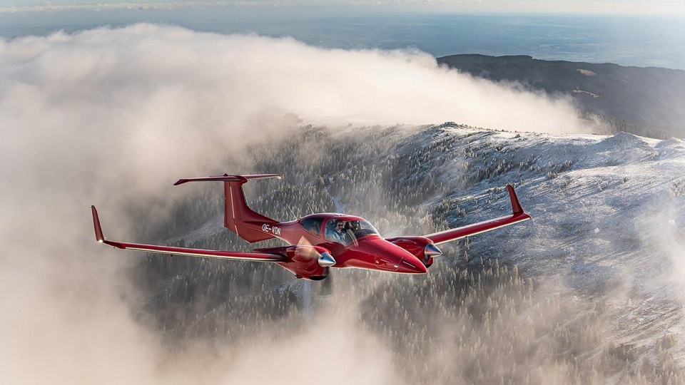 Diamond DA42-VI rouge et blanc en vol au-dessus d'une mer de nuages et de montagnes enneigées, vue trois-quarts avant